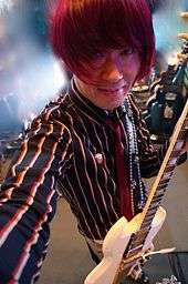 A picture of a man with dark pink hair, holding a guitar.