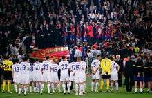 A line of men in white shirts watching men in blue-and-red shirts standing on a rostrum in the background.