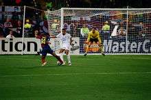 A man in a blue-and-red shirt with a football at his feet runs at a man in a white shirt. In the background, a man in a yellow shirt prepares for the ball to be kicked towards him.