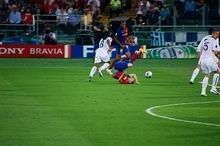 A man in a blue-and-red shirt is lying on the floor with his legs above his head. Behind him, a man in a white shirt is chasing after a football.