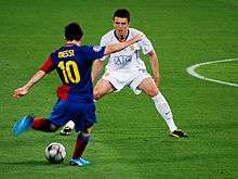 A man wearing a blue-and-red football shirt prepares to kick a football, while a man in a white shirt looks on.