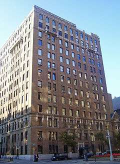 A twelve-story brown brick building, lit from the left by late afternoon sun, in front of an intersection with a traffic light