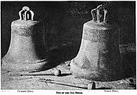 Two large church bells resting on a stone pavement, each carved with a indistinguishable inscription.