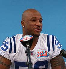 African American male in football uniform seated at a press conference with Super Bowl XLI logo in the background and an NFL logo on the microphone