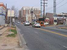 Ground-level view of a mid-sized street in an urban area, with one lane closed in the direction headed away from the viewer. In the far background are high-rise office and residential buildings, while the near background features a construction site with a large crane against an overcast sky on a winter day.