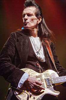 The head and torso of a middle-aged man. He has long, dark hair. He is wearing a dark jacket, a light top, and several items of jewellry, including large earrings. He is playing an electric guitar.