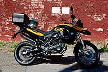 Black and yellow BMW F800GS with topbox and motorcycle helmet on the seat, parked in front of a brick wall covered in peeling red paint