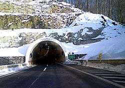 Tunnel portal in a snow covered slope, traffic sign indicating name of the tunnel is visible to the right of the entrance to the tunnel