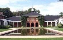 A red brick building and three sides of a white columned cloister behind a large reflecting pool