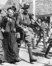 Troops marching through a city street lined with a crowd of people. In the foreground is a soldier and beside him is an older women.