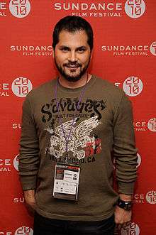 Color half-length portrait of Green: smiling man in jumper standing by red Sundance wall.