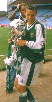 A brown-haired footballer holding a silver trophy adorned with green ribbons. He is wearing a green jersey with white sleeves, and white shorts.