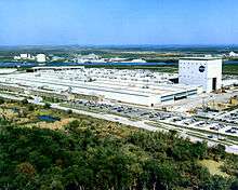 An aerial view of a large white building with green trees in the foreground and a blue sky in the background