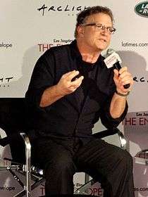 A white male sitting down while speaking into a microphone that he is holding with his left hand