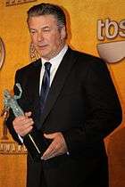 A man in his early fifties holds an awards while he is being photographed. He wears a dark tuxedo.