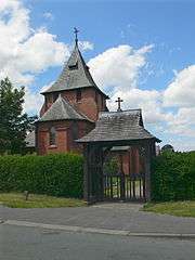 A lych gate with a hedge on each side; behind is the end of a polygonal apse with a pointed roof and beyond that a tower with a broach spire