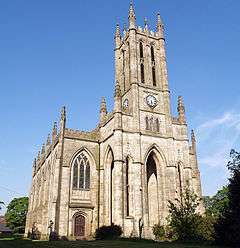 A large church seen from the northwest, showing a tower with clock faces over arches, an arched window, and crocketted pinnacles.