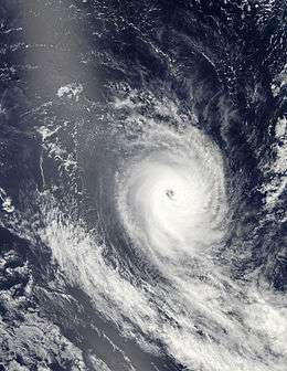 Satellite image of a tropical cyclone with an eye and spiraling rainbands. A small, green island is visible to the left of the cyclone.