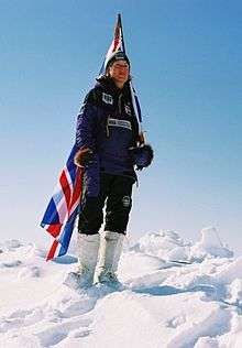 A figure standing on snow in dark clothing and white boots carries a British flag.