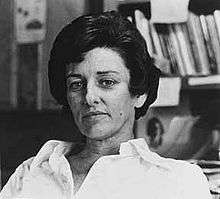 Head and shoulders monochrome portrait photo of Anne Sexton, seated with books in the background