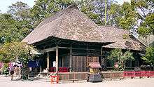 Three-quarter view of a wooden building with a thatched hip roof. Another structure of similar style extends from the back of the building.