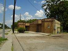 A one-story tan colored building. Bushes, trees, a sidewalk and a sky with multiple clouds surround the building.