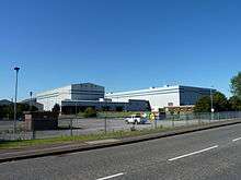 A photograph of a modern factory with two large industrial buildings standing side by side and a loading area at the front. The name of the company is written in red lettering at the front of each building. A colummn of steam rises in the background