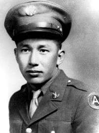 A young man in his army dress uniform. He is sitting and staring forward towards the camera with and he is not smiling. He has a high brimmed hat.