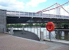 Start of a footbridge that curves under one end of a suspension bridge, along the bank of a wide river.