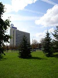 Long view of a light colored multistory factory through trees