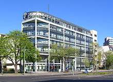 View of a seven-story, modern building, predominantly grey and white, with a cross-like symbol and large letters spelling "Scientology Kirche" at the top.