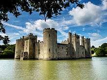 Photo of Bodiam Castle across the water, framed by oak leaves. The alternation of large round towers at the corners and square towers central to each face can be seen. Daylight visible through upper windows indicates that the roofs and inner structures are ruined.