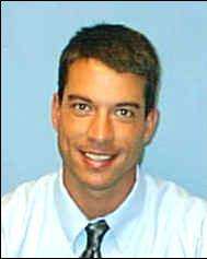 A smiling clean-shaven young Caucasian man with short dark hair wearing a white shirt and silvery tie against a blue background