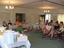 Women sitting in chairs at a party, with a few gifts stacked on a table in the foreground.