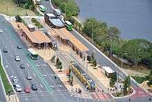 An aerial view of the station and bus interchange. The station is a single track station with platforms on either side. There are street-level open-air structures featuring passenger canopies on both platforms. Bus stops and roads are visible either side of the station.