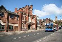 modern street scene, of broadgate with a bus heading toward us.  But on the far side of the road is the astonishing mock-gothic frontage of the Drill Hall, in redbrick with stone details, including two turret towers, pierced with cruciform windows