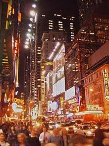 Photograph of a crowded city street at night. The street is in a commercial district; the buildings all have at least several stories, and some are high rise. The buildings have elaborate signs, many of which incorporate neon lighting. There are prominent signs for Madame Tussaud's, Loew's, Empire, AMC 25 Theatre, and Modell's.