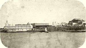 Photograph depicting a waterfront with stone quays, a paved ramp leading down to the water's edge, a fortress on the left edge of the picture, and spires, domes and multi-story buildings in the background