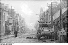 A soldier walks up a high street; the sides of the road are covered with rubble. In the foreground there is a knocked out tank, at the foot of the tank there is a dummy wooden gun barrel and debris. Another knocked out tank being examined by another solder occupies the background.