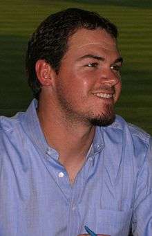 A smiling dark-haired young man with a dark goatee wearing a blue button-down shirt