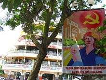 A poster showing a red sky emblazoned with the hammer-and-sickle above a stylized city landscape. In the foreground stands a light-skinned man wearing an orange hat, a blue collared shirt and cyan gloves; he holds a paper with a symbol of atomic energy in his right hand. At the foot of the poster is Vietnamese text. Behind the poster is a large, white, multi-storey building behind a tree.