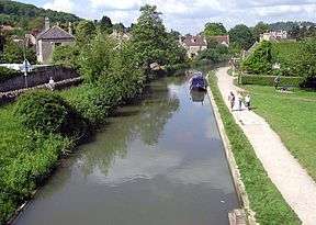 Wide stretch of still water with a narrow boat on it. Tow path on the right and trees and houses on the left.