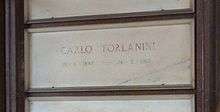 A white marble gravestone on the wall of a chapel, with only the name and dates of birth and death inscribed