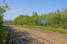 A muddy road with deep tire ruts and puddles on the left, amid a wooded area. At the left is a white on green sign with "Winter Ice ... Maximum Weight ... 000 kg" written on it. In the distance is some water reflecting evergreen trees across it