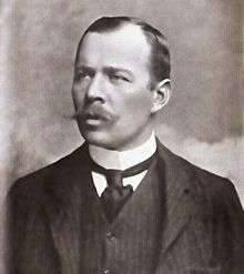  Head and shoulders portrait of a man with receding hair, heavy moustache, looking left from the image. He wears a high white collar, black necktie, dark waistcoat and jacket.