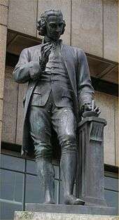 Statue of a man with a mortar and pestle in his left hand and his right-hand upraised.