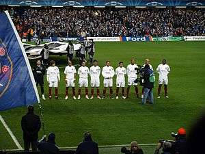 group of several men in several rows, standing, dressed in black and white football kit, flanked by man with camera