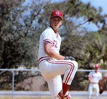 A man wearing a white baseball uniform and a red cap with a white "C" on it winds up to throw a baseball from the pitcher's mound