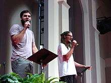 A photograph of two men with facial hair wearing t-shirts while speaking into black handheld microphones and gripping black music stands indoors