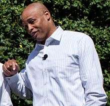 A man wearing a buttoned shirt stands in front of a vine and smiles.